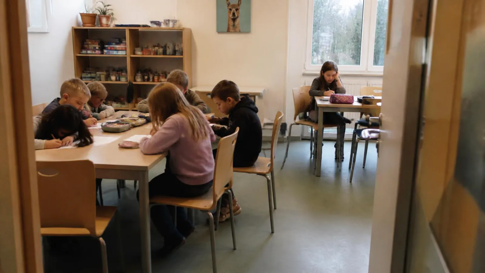 Illustration (KI-generiert): Kinder machen am großen Tisch Hausaufgaben, ein Mädchen sitzt allein am Fenster